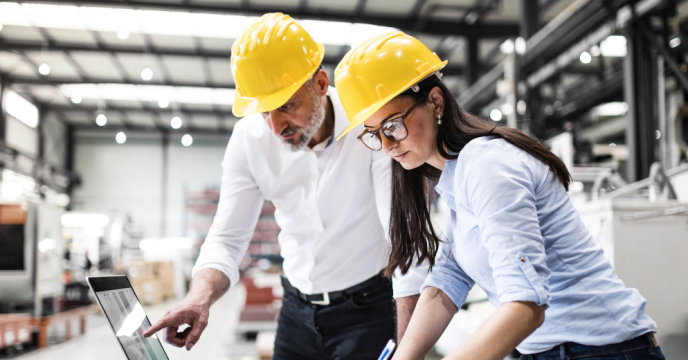 Two people wearing hard hats looking at laptop at manufacturing site